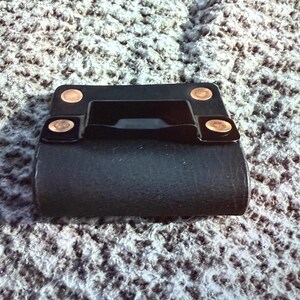 May include: A black leather belt loop with copper rivets. The loop has a rectangular shape with a curved bottom and a handle-like feature in the center. The rivets are circular and add a decorative touch.