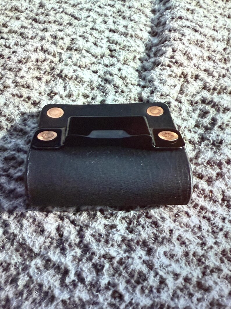 May include: A black leather belt loop with copper rivets. The loop has a rectangular shape with a curved bottom and a handle-like feature in the center. The rivets are circular and add a decorative touch.