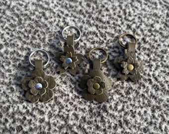 Op de afbeelding: Vier olijfgroene leren bloemvormige sleutelhangers met zilveren en messing sleutelringen. Elke bloem heeft een ander middendetail, inclusief zilveren en messing studs. Op een getextureerd, neutraal gekleurd oppervlak.