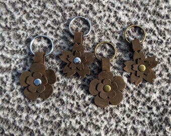 Op de afbeelding: Vier bruinleren bloemen sleutelhangers met metalen sleutelringen. Elke bloem heeft een ander midden: zilver, zilver, goud en goud. De sleutelhangers zijn gerangschikt op een getextureerd, grijs oppervlak.