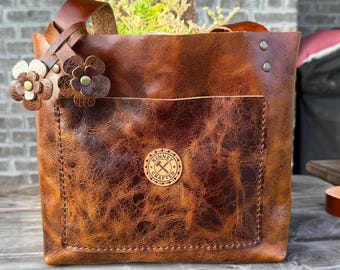 Op de afbeelding: Een bruine leren tas met een getextureerde afwerking, voorzien van een voorvak, twee leren bloemhangers bevestigd aan de riem en een ronde logopatch met de tekst Kinney Crafted en een icoon van een hamer en moersleutel op het voorvak.