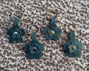 Op de afbeelding: Vier teal leren bloemvormige sleutelhangers met zilver- en goudkleurige metalen ringen. Elke bloem heeft een gelaagd bloemblaadjesontwerp en een centrale stud. Gepresenteerd op een getextureerd, neutraal getint oppervlak.