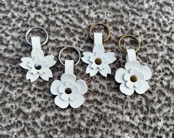 Op de afbeelding: Vier witte leren bloemvormige sleutelhangers met metalen ringen. Elke bloem heeft een ander middelpunt: een zilveren, een zwarte en twee bronzen accenten. De sleutelhangers zijn gerangschikt op een getextureerd grijs oppervlak.