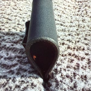 May include: A black, cylindrical object with a textured surface. The object has a flared, teardrop-shaped opening at the bottom and a small handle-like projection on the side. The interior is dark, and the object appears to be made of a rigid material.