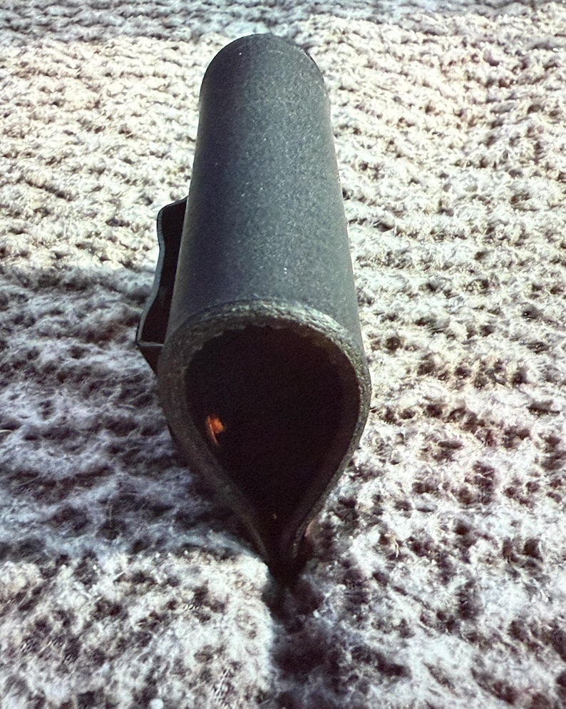 May include: A black, cylindrical object with a textured surface. The object has a flared, teardrop-shaped opening at the bottom and a small handle-like projection on the side. The interior is dark, and the object appears to be made of a rigid material.