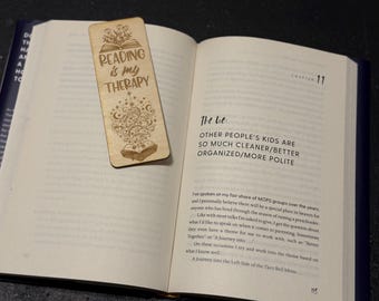 Op de afbeelding: Een lichtbruine houten bladwijzer met de tekst "Reading is my Therapy" en decoratieve illustraties, liggend op de open pagina's van een boek. De bladwijzer heeft geëtste ontwerpen.