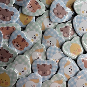 Könnte beinhalten: Eine Sammlung herzförmiger Anstecknadeln mit Cartoon-Tierdesigns. Die Anstecknadeln haben ein Vichy-Muster in Pastellfarben mit Bildern von Bären, Hasen und Küken. Einige Anstecknadeln tragen Namen.