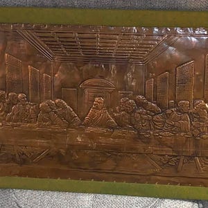 May include: A rectangular copper wall plaque featuring a detailed embossed relief of The Last Supper. The metallic artwork depicts a central figure seated at a long table with twelve individuals, framed by a solid olive green border on all four sides.