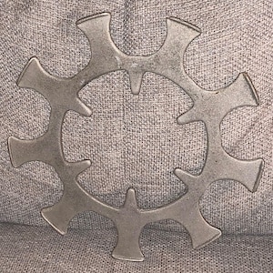 May include: A silver-colored, circular metal trivet with a unique, openwork design. The trivet features a series of stylized, curved arms radiating from a central ring, creating a decorative and functional piece for protecting surfaces from hot items.
