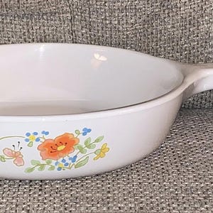 May include: White, oval-shaped skillet with a single handle. The skillet features a floral design with orange, blue, and yellow flowers, and a butterfly. The skillet is made of a durable material, perfect for cooking.