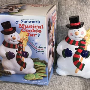 May include: A white ceramic snowman cookie jar with a black top hat, red and green scarf, and broom. The jar is next to its box, which reads "Snowman Musical Cookie Jar". The jar holds 1.5 pounds of cookies.