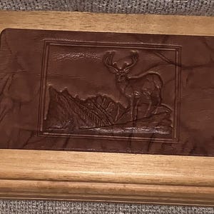 May include: A rectangular wooden box with a brown leather inset. The inset features a detailed relief of a deer standing in a mountainous landscape. The wood frame is light brown.