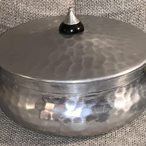May include: A silver hammered aluminum pot with a lid and black handles. The lid has a black knob on top.