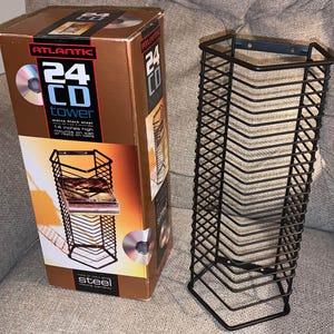 May include: A matte black steel Atlantic 24 CD tower storage rack with a grid design. The packaging box displays the text 24 CD TOWER, matte black steel, and 14 inches high, mounts on wall or rests on table, showing the rack's vertical structure.