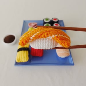 May include: A crocheted sushi set with a variety of pieces, including a yellow and black nigiri, a red and white nigiri, a green and white nigiri, and a yellow and orange maki roll. The sushi is displayed on a blue tray.
