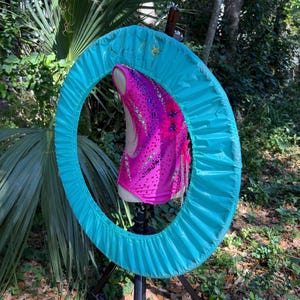 以下が含まれることがあります： ターコイズ色の布製のフープの中に、ラインストーンとスパンコールがついたピンクとパープルのレオタードが入っています。レオタードはマネキンに着せられています。