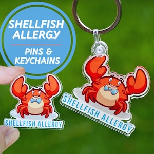 May include: A blue circle with the text "Shellfish Allergy Pins & Keychains". A red cartoon crab with a blue bow tie is on a white background with the text "Shellfish Allergy".