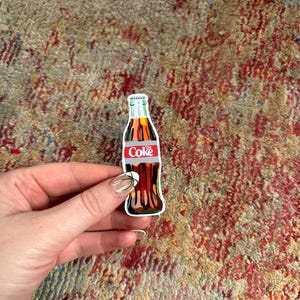 May include: A vintage-style sticker of a glass bottle of Coca-Cola. The bottle is brown and has a red label with the word "Coke" in white letters.