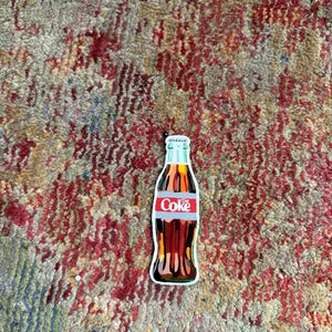May include: A vintage-style Coca-Cola bottle illustration with a red and white label that says "Coke". The bottle is brown and has a white cap. The illustration is on a white background.