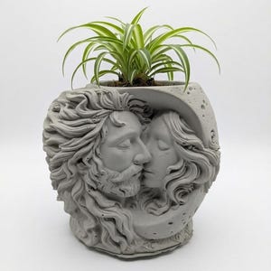 Könnte beinhalten: Ein hellgrauer Betonübertopf mit einem skulpturalen Relief eines bärtigen Gesichts und eines Profils, die in eine Mondsichelform eingebettet sind. Der Übertopf enthält eine kleine grün-weiße Grünlilie mit langen, dünnen und spitzen Blättern.