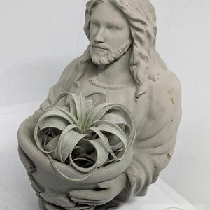 May include: A gray concrete statue of a figure holding a bowl with a plant. The statue has long hair and a beard. The plant has long, curled leaves. The statue is a decorative item.