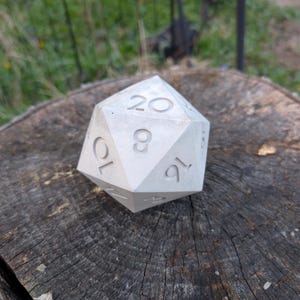 May include: A white, 20-sided die with numbers 1 through 20 etched on each face. The die is made of concrete and is sitting on a wooden surface.