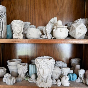 May include: A collection of handmade, light gray concrete planters and decorative objects. The shelf displays various face planters, a D20 die, a small pot with a lid, and other unique sculpted items. The items are arranged on wooden shelves.