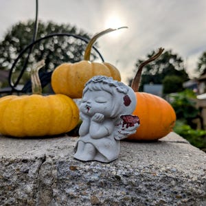 May include: A small, gray angel figurine with red accents sits in front of three pumpkins. The pumpkins are various shades of orange and yellow. The scene is set outdoors with a cloudy sky in the background.
