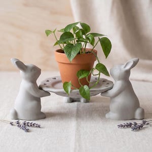 May include: Two light gray rabbit figurines standing on their hind legs, supporting a circular gray plate with a heart-patterned rim. A small terracotta pot containing a green leafy plant sits centered on the plate, flanked by sprigs of dried lavender.