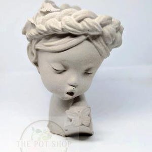 May include: A light beige ceramic planter shaped like a young person's head, with closed eyes and a braided hair crown. The figure holds a butterfly. The Pot Shop logo is at the bottom.