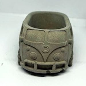 May include: A small, gray concrete planter shaped like a vintage Volkswagen van. The planter has a rounded, rectangular shape and features a detailed design of the van's front, including the headlights, grille, and iconic VW logo.