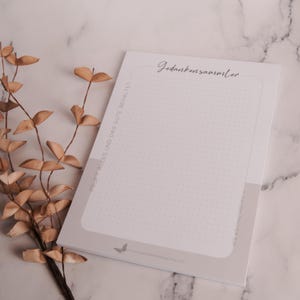 May include: A white notepad with a dotted grid pattern and the German text "Gedankensammler" in elegant script. The notepad has a light grey border and a butterfly graphic. Dried leaves are in the background.