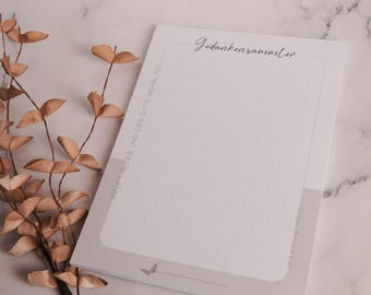 Puede incluir: Un bloc de notas blanco con un patrón de cuadrícula punteada y el texto alemán "Gedankensammler" en escritura elegante. El bloc de notas tiene un borde gris claro y un gráfico de mariposa. Hojas secas en el fondo.