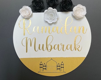 May include: Circular Ramadan Mubarak sign with gold lettering and a mosque silhouette. The sign is white with a gold lower section and is decorated with black and white paper flowers. A white rope is attached for hanging.
