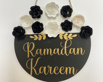 May include: A circular Ramadan Kareem sign in black and white, featuring gold lettering and floral details. It's adorned with black and white paper flowers and suspended by a natural rope.