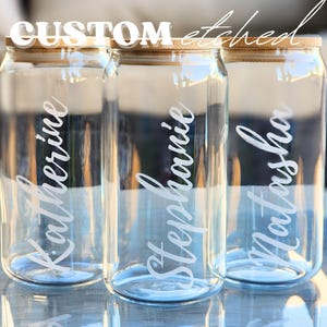 Op de afbeelding: Drie heldere glazen drankblikken met bamboe deksels, elk geëtst met een andere naam in een cursief lettertype: Katherine, Stephanie en Natasha. De woorden "CUSTOM etched" staan boven de blikken.