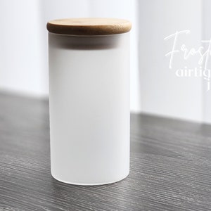 Może przedstawiać: Cylindryczny słoik z matowego szkła z jasnobrązową bambusową pokrywką. Napis "Frosted airtight jar" jest nadrukowany na biało po prawej stronie słoika. Pojemnik ma gładkie, półprzezroczyste wykończenie oraz płaską, okrągłą drewnianą pokrywkę.
