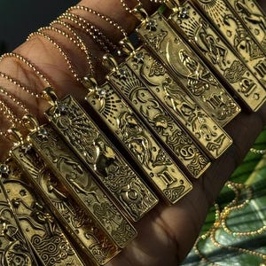 May include: A collection of gold-colored rectangular pendant necklaces, each featuring a different zodiac symbol. The pendants are suspended from delicate gold chains. The zodiac symbols are intricately detailed and embossed on the pendants.