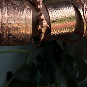 May include: Copper-coloured bracelets with detailed patterns. One has a hammered finish, another an embossed floral design. Two elephant head charms decorate a smooth copper bracelet. The bracelets are shown against a background of green leaves.
