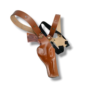 Op de afbeelding: Een bruin leren schouderholster met een bijpassende bruine leren riem en een zwarte elastische riem. Het holster is ontworpen om een ​​handvuurwapen te bevatten, en de houten handgreep van het pistool is zichtbaar.