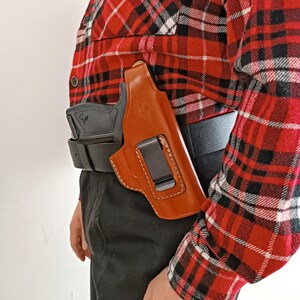Op de afbeelding: Een bruin leren holster met een zwarte clip is bevestigd aan een zwarte riem. Een zwart vuurwapen is gedeeltelijk zichtbaar in het holster. De persoon draagt een rood, zwart en wit geruit overhemd en een zwarte broek.