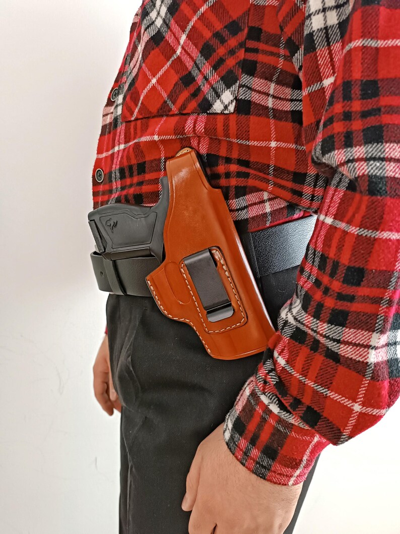 Op de afbeelding: Een bruin leren holster met een zwarte clip is bevestigd aan een zwarte riem. Een zwart vuurwapen is gedeeltelijk zichtbaar in het holster. De persoon draagt een rood, zwart en wit geruit overhemd en een zwarte broek.