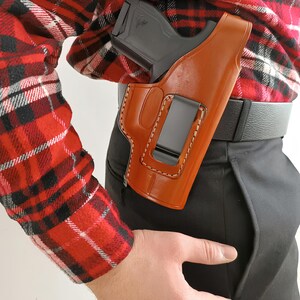 Op de afbeelding: Een bruin leren holster met een zwarte clip is bevestigd aan een zwarte riem. Een zwart pistool is gedeeltelijk zichtbaar in het holster. De persoon draagt een rood-zwart geruit overhemd en een zwarte broek.