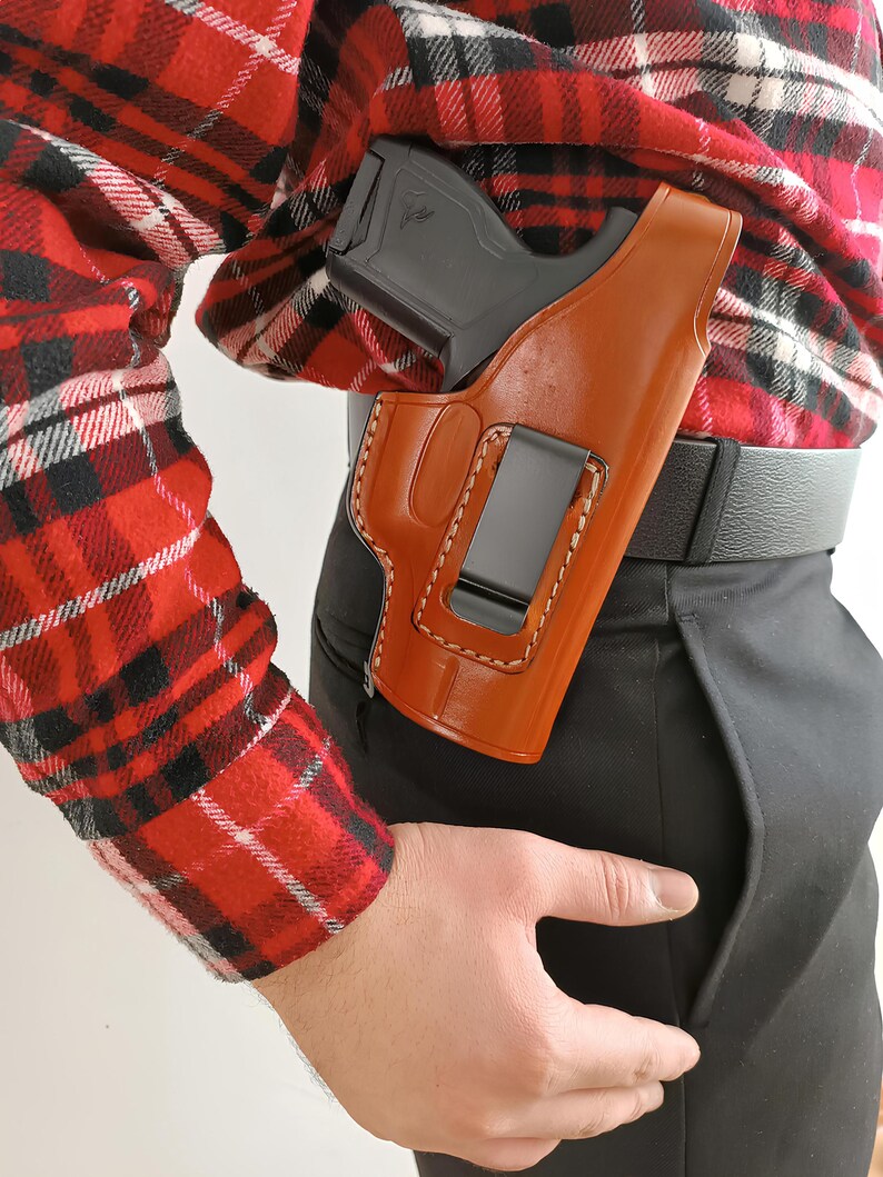 Op de afbeelding: Een bruin leren holster met een zwarte clip is bevestigd aan een zwarte riem. Een zwart pistool is gedeeltelijk zichtbaar in het holster. De persoon draagt een rood-zwart geruit overhemd en een zwarte broek.