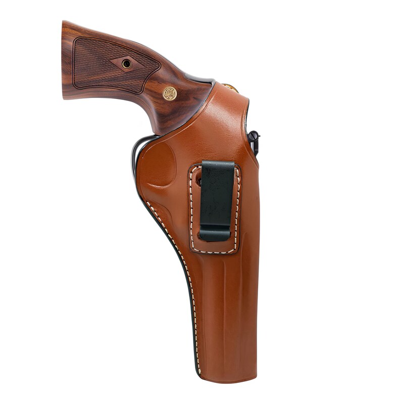 Op de afbeelding: Een bruin leren holster met witte stiksels en een zwarte clip. Het holster bevat een revolver met een houten handvat. De revolver heeft een gouden embleem aan de zijkant. Het holster is ontworpen om op de riem te dragen.