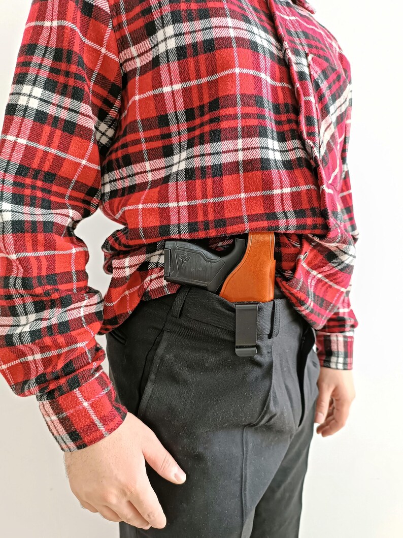 K&ouml;nnte beinhalten: Ein schwarzes und braunes Holster mit einer schwarzen Pistole ist am Bund einer schwarzen Hose befestigt. Die Person tr&auml;gt ein rot-schwarz-wei&szlig; kariertes Flanellhemd.