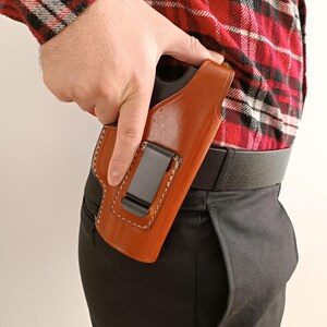 K&ouml;nnte beinhalten: Eine braune Lederholster mit wei&szlig;en N&auml;hten und einem schwarzen Metallclip. Die Holster ist f&uuml;r das Tragen im Hosenbund konzipiert. Die Person tr&auml;gt ein rot-schwarz kariertes Hemd und eine schwarze Hose.