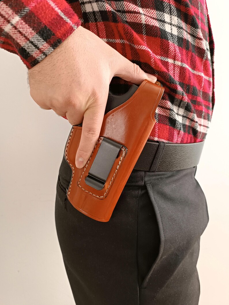 K&ouml;nnte beinhalten: Eine braune Lederholster mit wei&szlig;en N&auml;hten und einem schwarzen Metallclip. Die Holster ist f&uuml;r das Tragen im Hosenbund konzipiert. Die Person tr&auml;gt ein rot-schwarz kariertes Hemd und eine schwarze Hose.