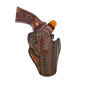 Op de afbeelding: Een bruin leren holster met een geweven mandweefpatroon, met een revolver met een houten handvat. De holster heeft oranje accenten en zwarte stiksels. De greep van de revolver is donkerbruin met een gouden embleem.