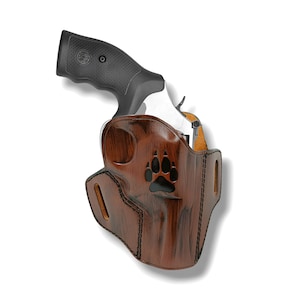 May include: A brown leather holster with a black paw print design, holding a silver and black revolver. The holster has a textured finish and is stitched with black thread. The revolver's grip is black.